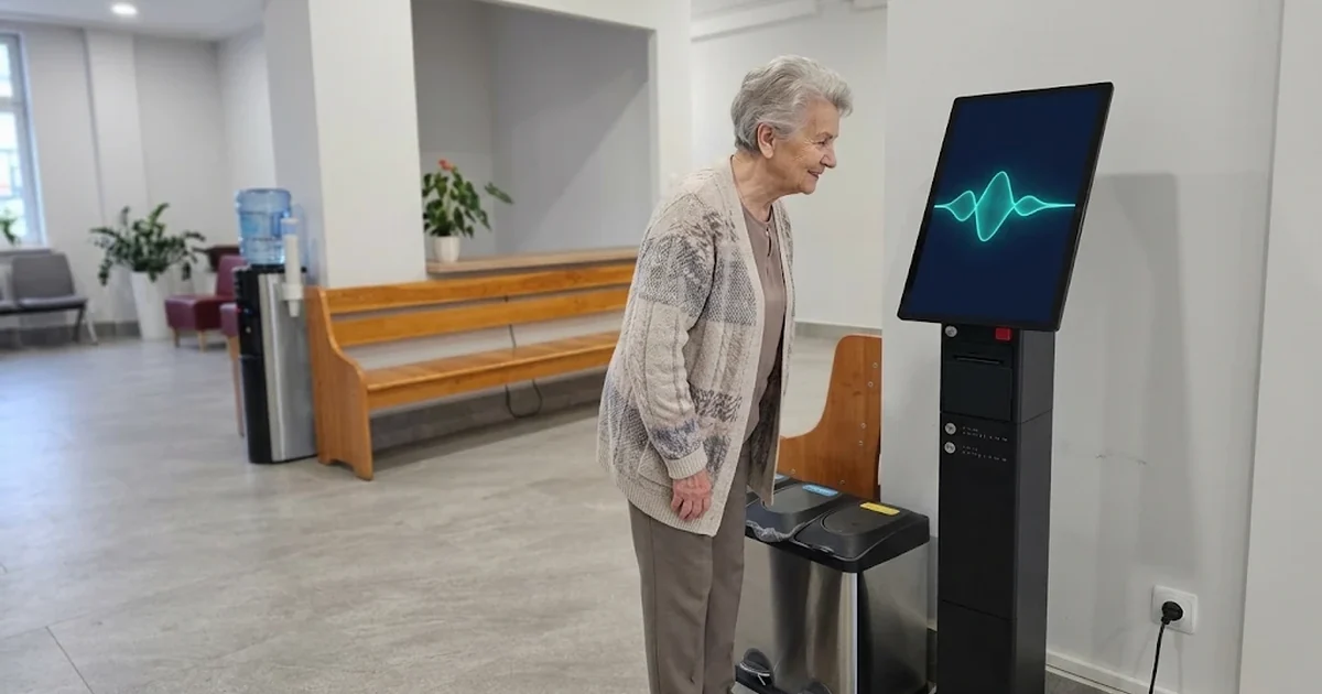 Starsza kobieta rozmawia z automatem biletowym QWinTech w holu instytucji; na ekranie automatu turkusowa fala dźwiękowa